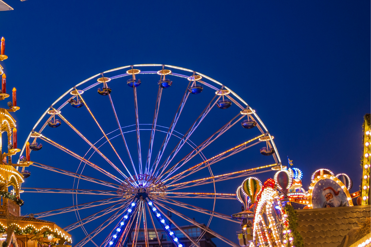L'immagine rappresenta una prospettiva dal basso della ruota panoramica con ai lati delle parti di bancarelle con decorazioni natalizie